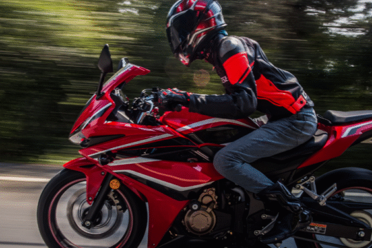 A motorcyclist in red riding gear speeding on a red sports bike on a highway, with trees blurred in the background.