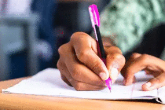 Student writing with a pink pen in a classroom notebook.