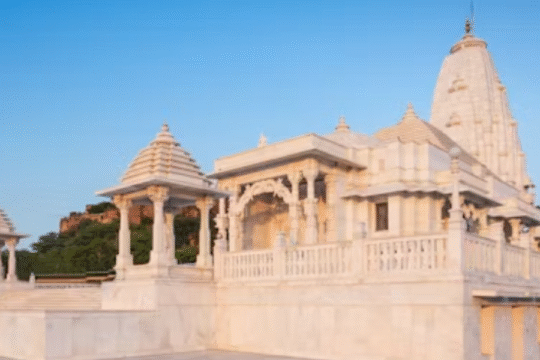 White marble Hindu temple with domes and pillars, surrounded by greenery under a clear blue sky.
