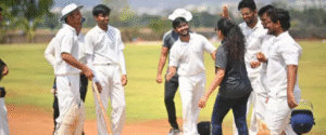 Group of young cricketers in white uniforms celebrating with a coach on a sunny day at a cricket ground.