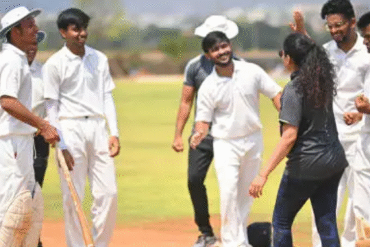 Group of young cricketers in white uniforms celebrating with a coach on a sunny day at a cricket ground.