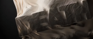 A mysterious draped object partially revealing bold block letters beneath a flowing beige fabric.