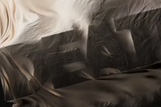 A mysterious draped object partially revealing bold block letters beneath a flowing beige fabric.