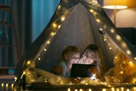 Mother and child watching tablet together inside a cozy tent decorated with string lights and stuffed toys.