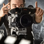 Film director framing a shot through a camera on a smoky movie set with lights and a clapperboard in the foreground.