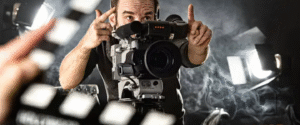 Film director framing a shot through a camera on a smoky movie set with lights and a clapperboard in the foreground.