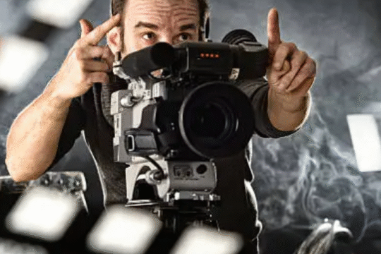 Film director framing a shot through a camera on a smoky movie set with lights and a clapperboard in the foreground.