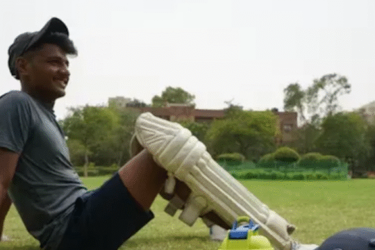 A boy sitting on a cricket ground, looking happy and tried, wearing full cricket gear.
