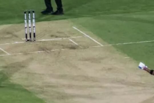 Indian and Australian cricketers on the pitch during a T20 match, with Virat Kohli raising his bat in celebration near the crease.