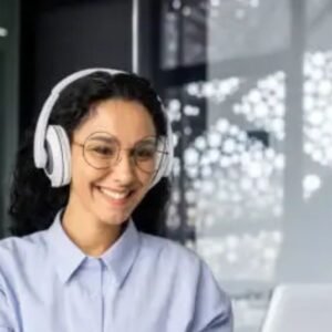 A smiling woman wearing headphones and glasses working on a laptop in a modern office setting, with papers and a pencil on the desk.