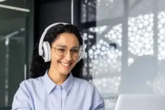 A smiling woman wearing headphones and glasses working on a laptop in a modern office setting, with papers and a pencil on the desk.
