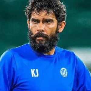 A man with a beard and curly hair in a blue shirt holds a soccer ball.