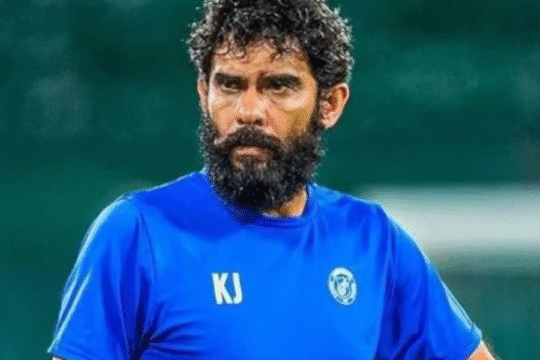 A man with a beard and curly hair in a blue shirt holds a soccer ball.