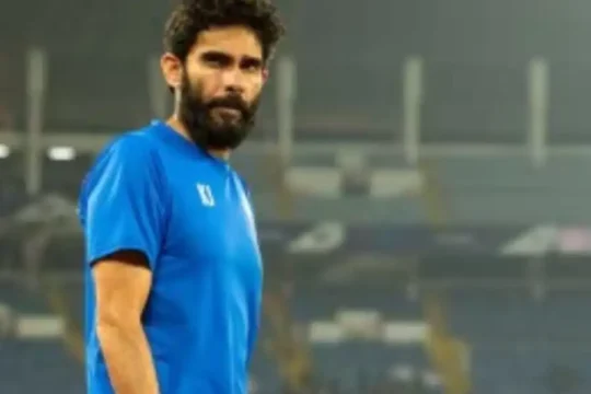 A serious bearded man in a blue shirt on a football field.