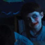 A young boy with pale makeup and red lipstick smiling creepily in a classroom.